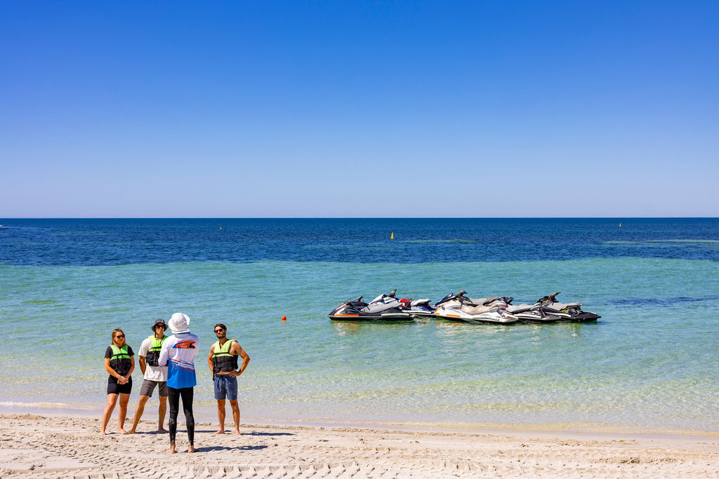 People in swimwear on a beach near jet skis and calm blue ocean.