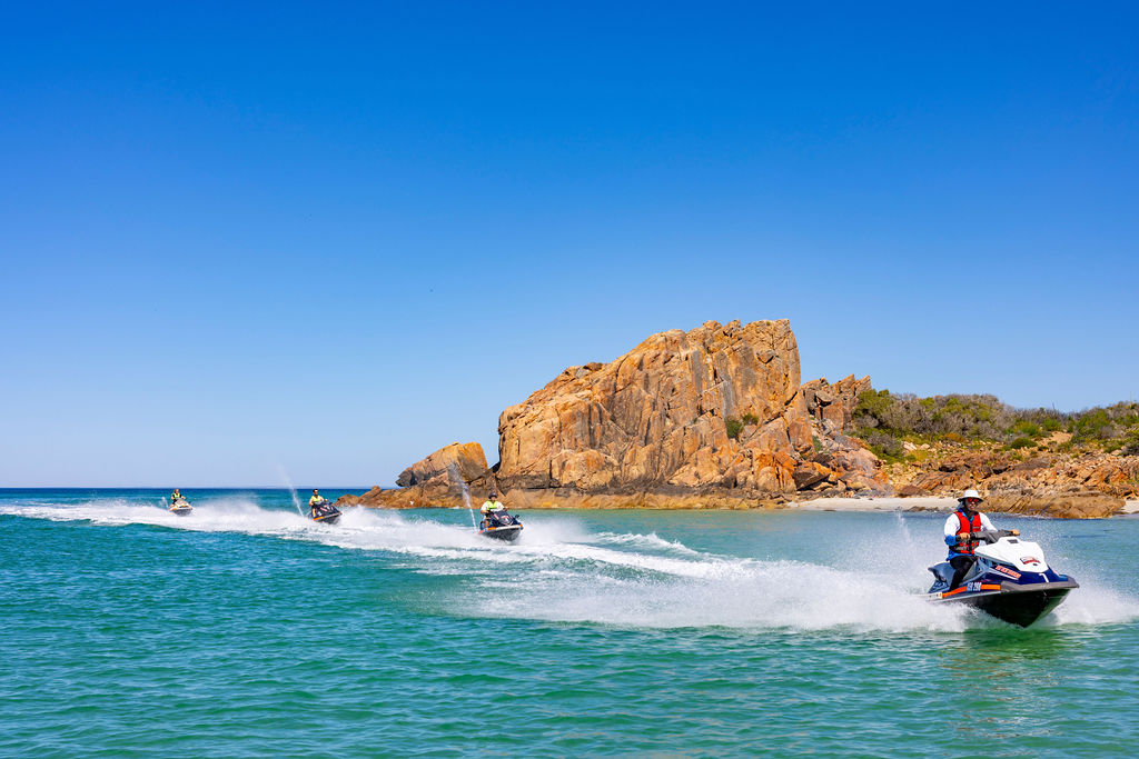 Four jet skis racing near a rocky shoreline on a clear, sunny day.