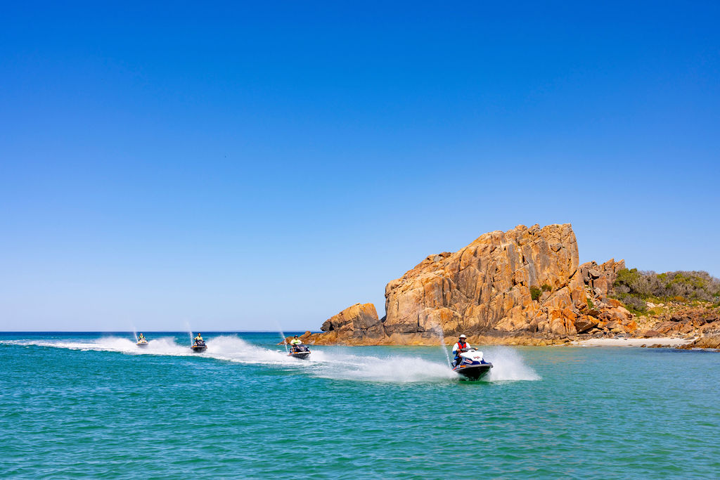 Four jet skis speeding on blue water near a rocky island.