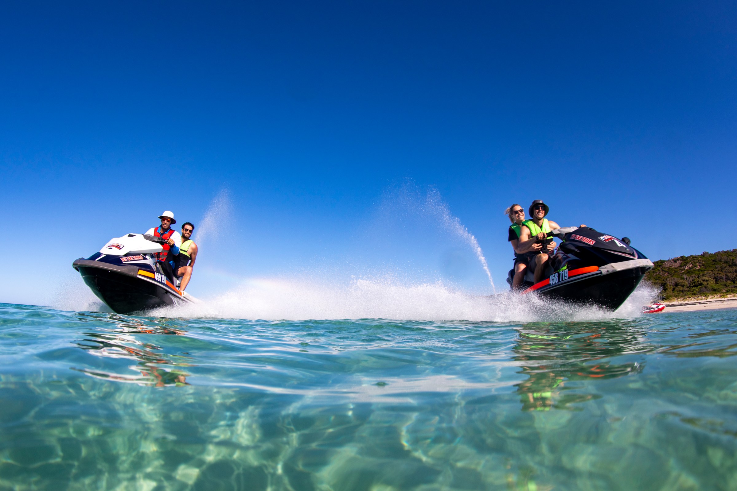 Two people on jet skis speeding over clear blue water under a bright sky.