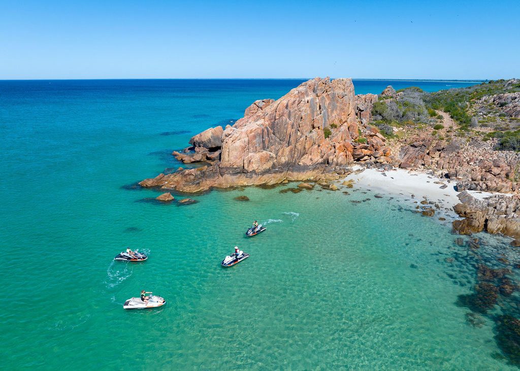 Jet skis near rocky coastline and sandy beach in turquoise water.