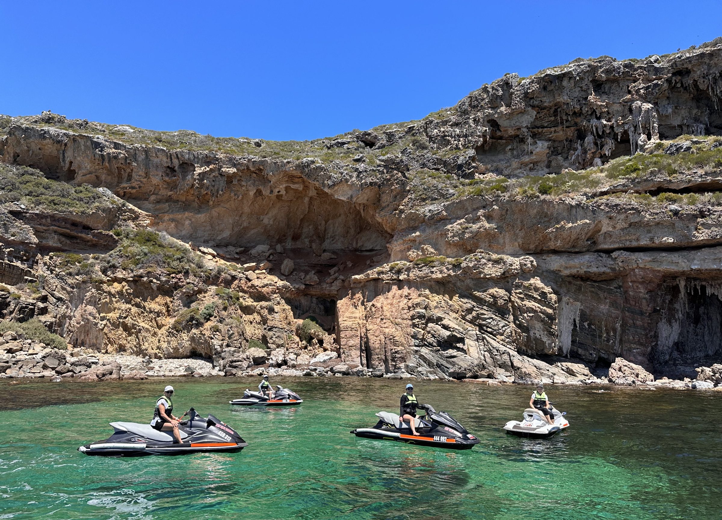 People on jet skis in clear water near rocky cliffs under a blue sky.